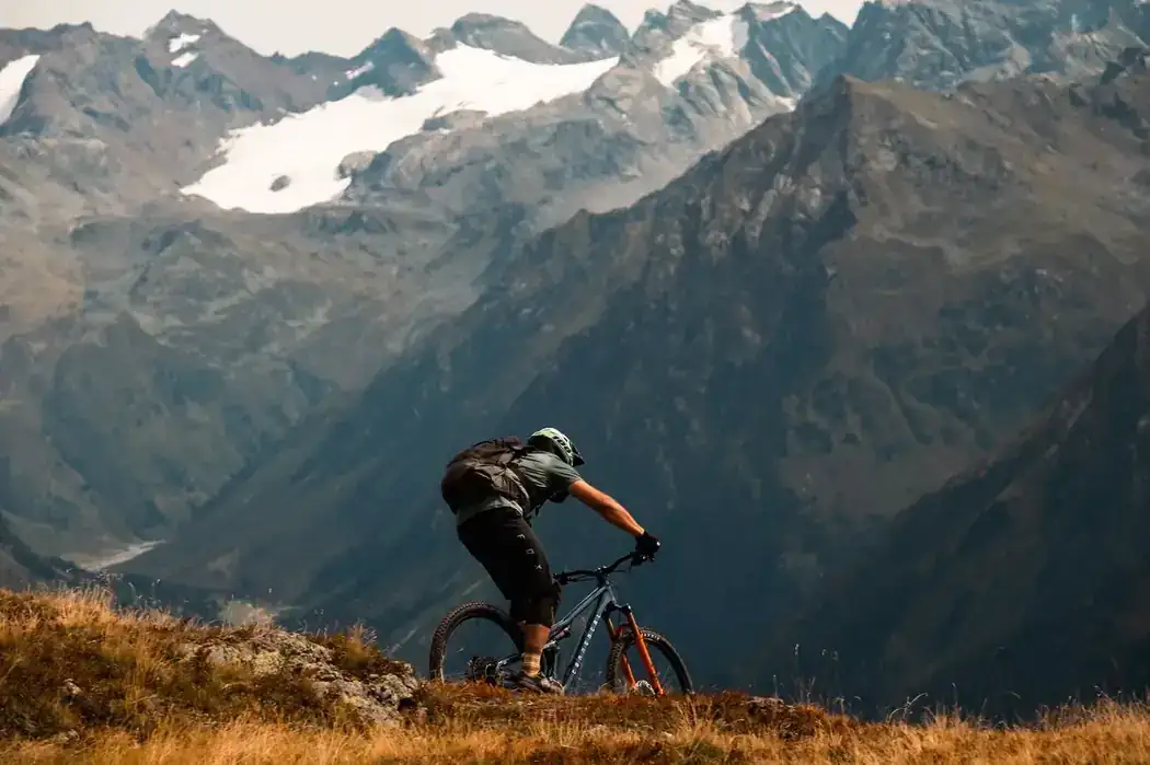 Mountain biker descending an alpine singletrack trail with snow-capped peaks in the background